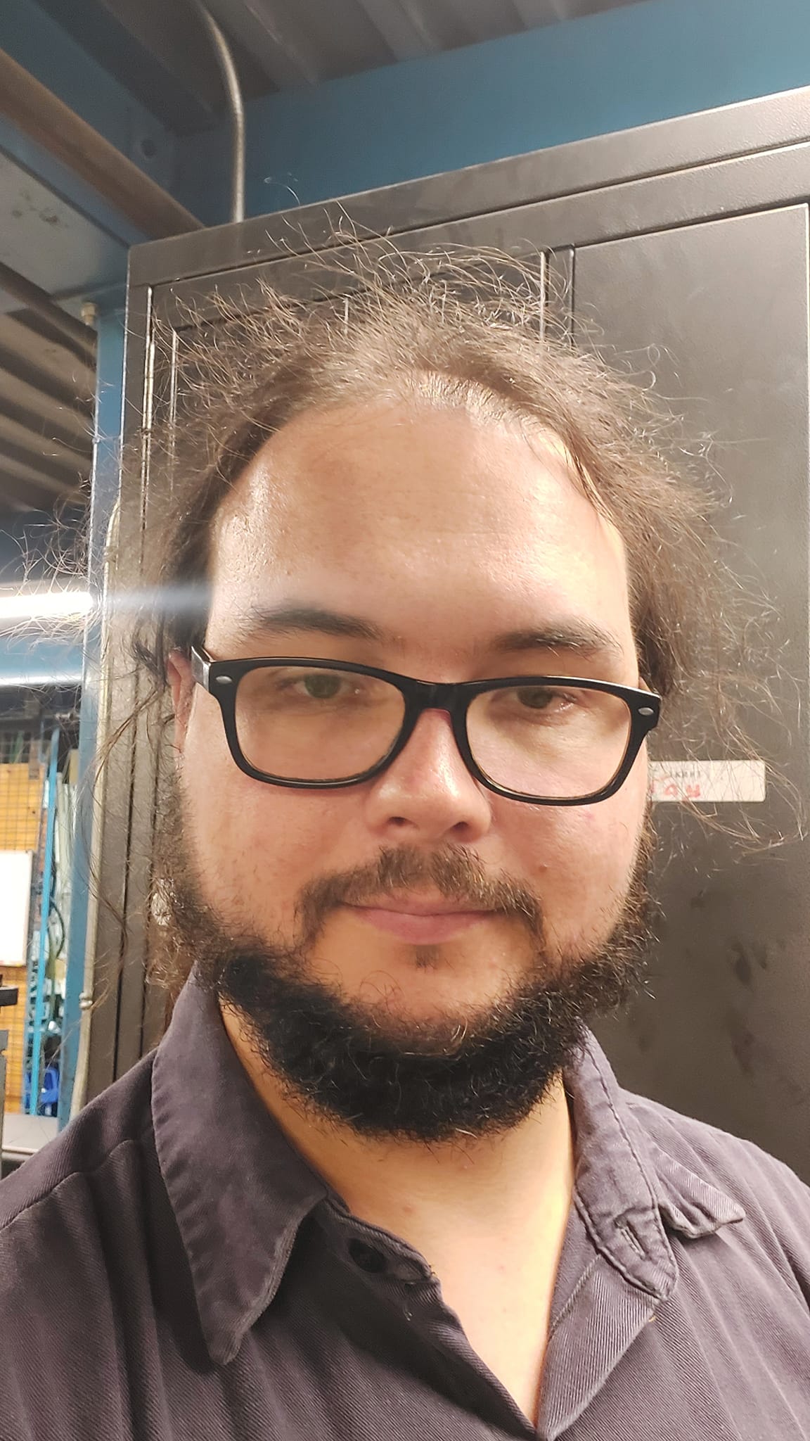 A man with tied back long black hair stands in an industrial maintenance office, photo 1